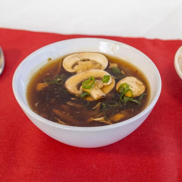 A white bowl of hot and sour soup with mushrooms and green onions on a red tablecloth.