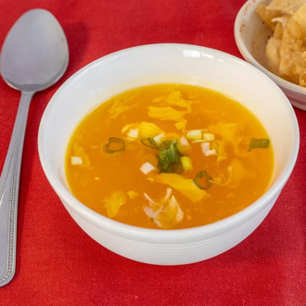 A white bowl of golden egg drop soup with green onions on a red tablecloth, with a spoon and fried wontons.