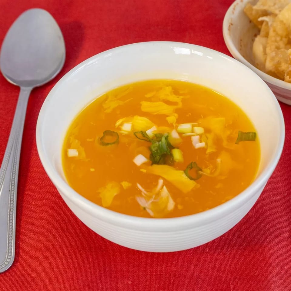 A white bowl of golden egg drop soup with green onions on a red tablecloth, with a spoon and fried wontons.
