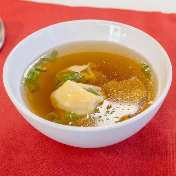 A white bowl of wonton soup with green onions on a red tablecloth at China Bell Restaurant.